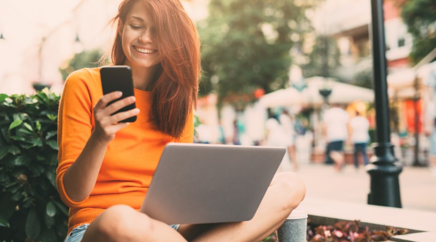 young woman using phone and laptop at an outdoor cafe